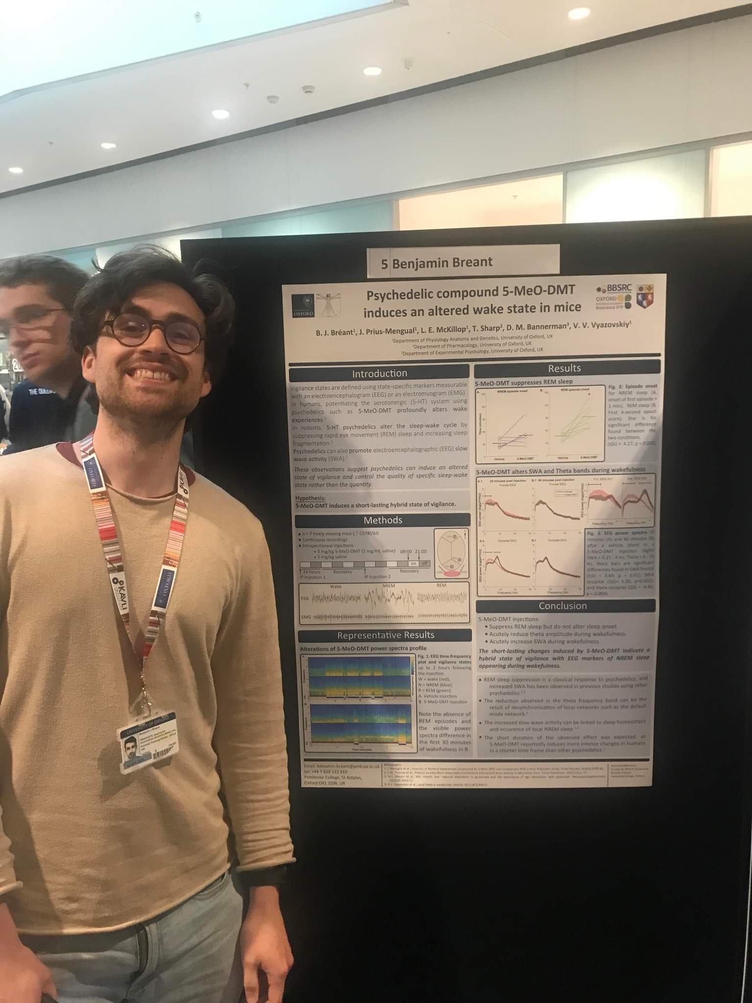 Benjamin Bréant standing in front of his academic poster that he presented at the Doctoral Training Centre. Ben is smiling, wearing a beige long sleeve shirt and his colourful lanyard.