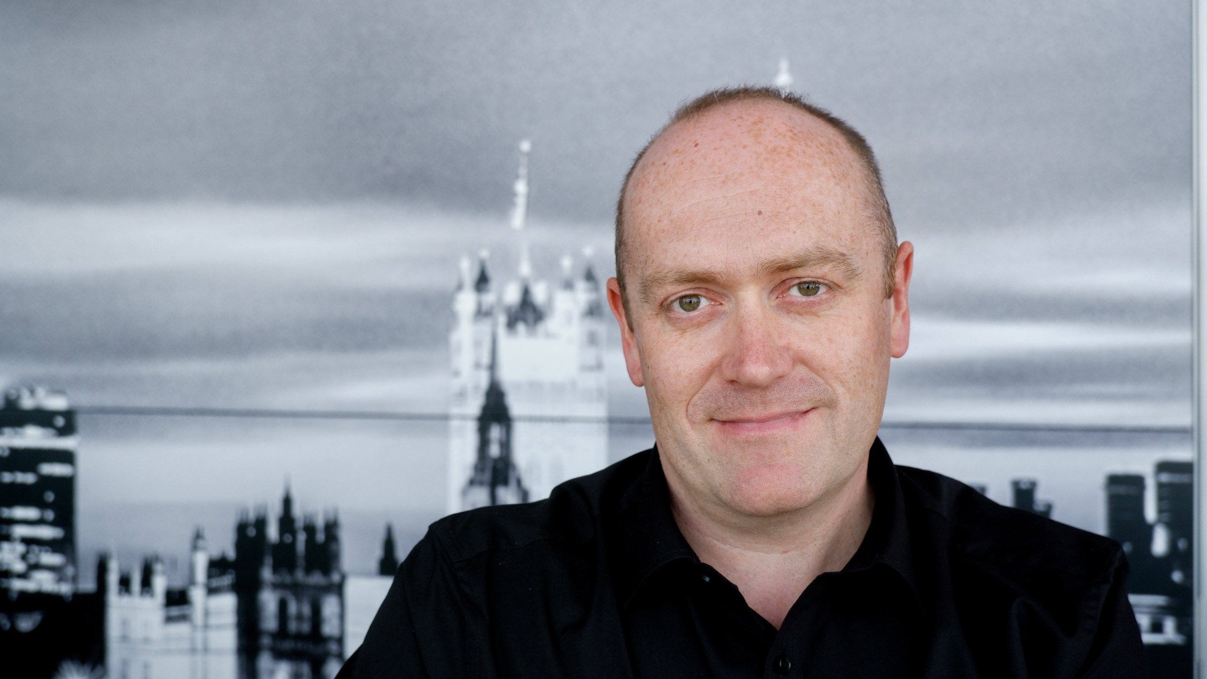 Headshot of Cennydd smiling, he is wearing a black shirt and a black and white image of a city is behind him