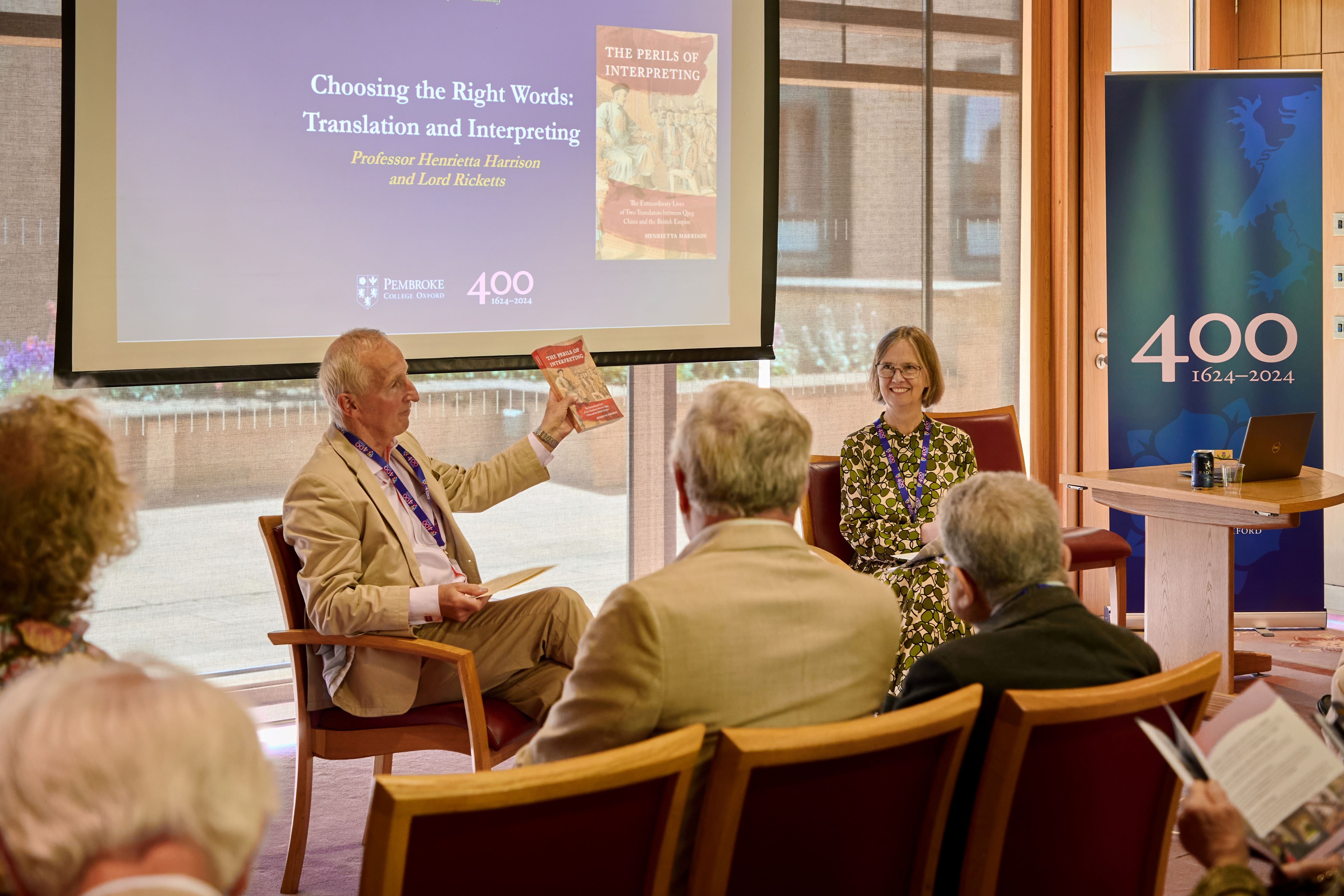 Lord Ricketts and Prof Henrietta Harrison sit on the panel for a speaker session. Lord Ricketts is holding a copy of Prof Harrison's book in the air.
