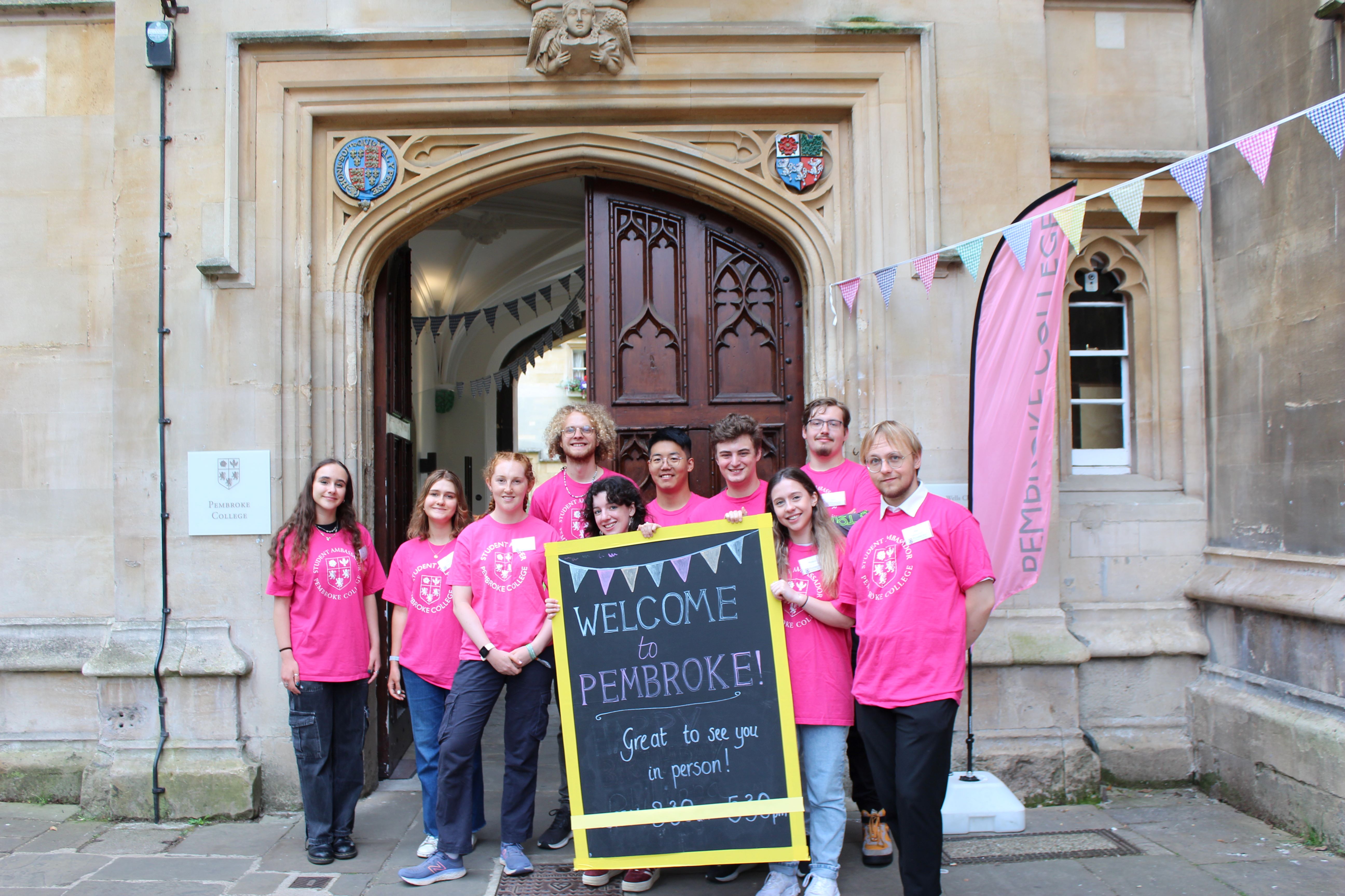 Group of student ambassadors standing outside Pembroke College in pink t-shirts. They are holding a sign that says "Welcome to Pembroke, Great to see you in person!"
