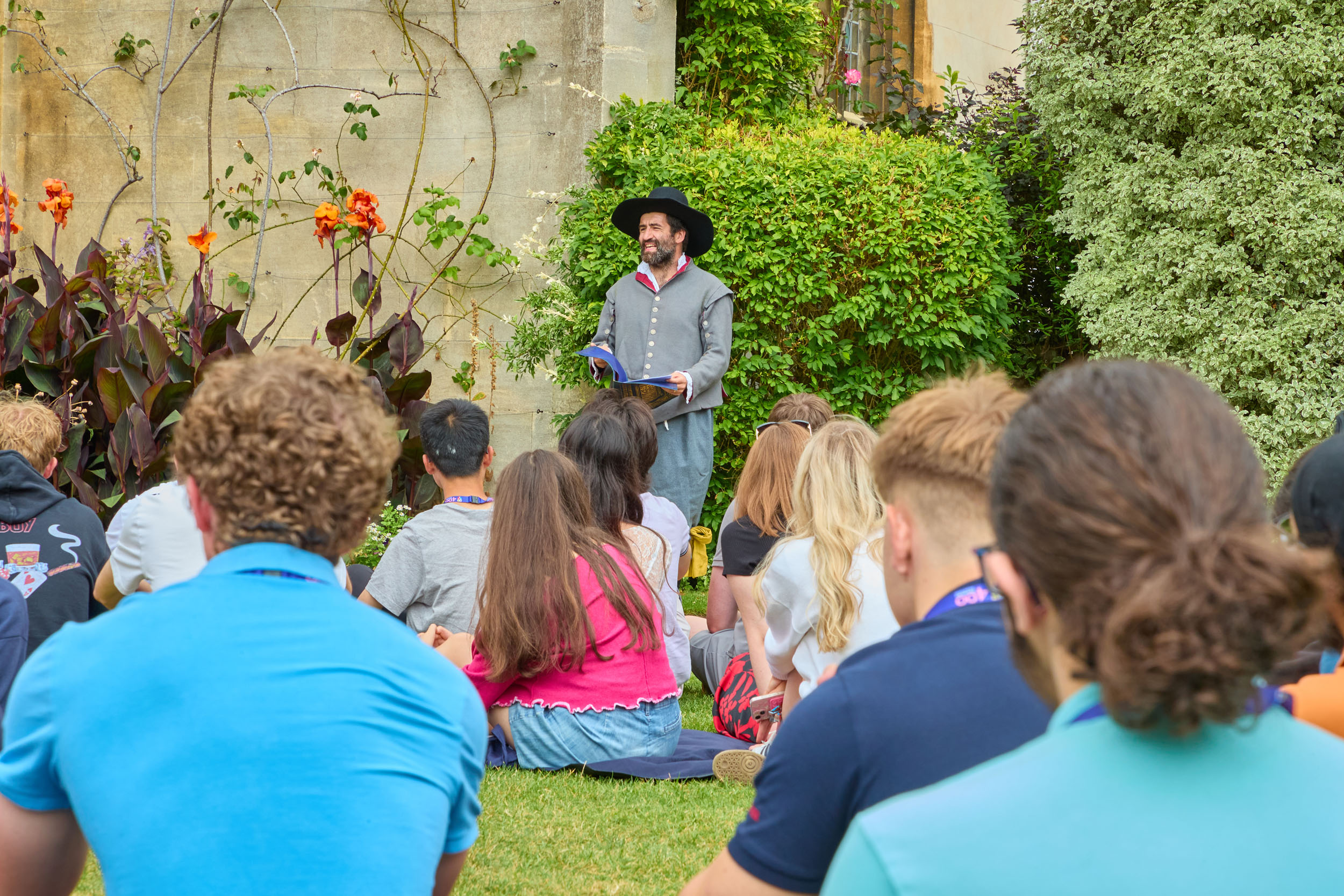 A man in 17th century dress reads from a book outside Broadgates Hall, with a crowd of students sat in front of him.