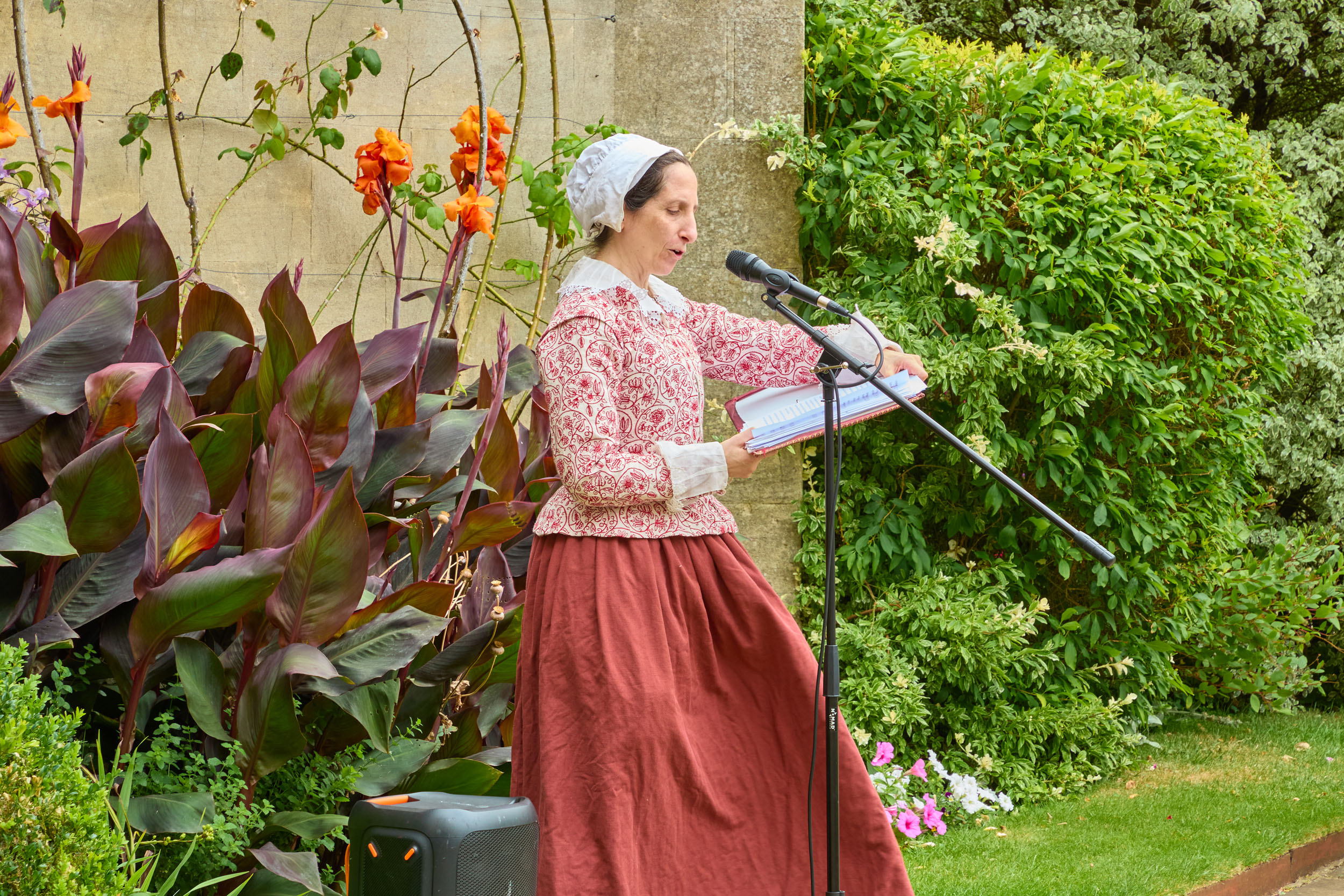 A woman in 17th century dress reads from a book in front of Broadgates Hall.