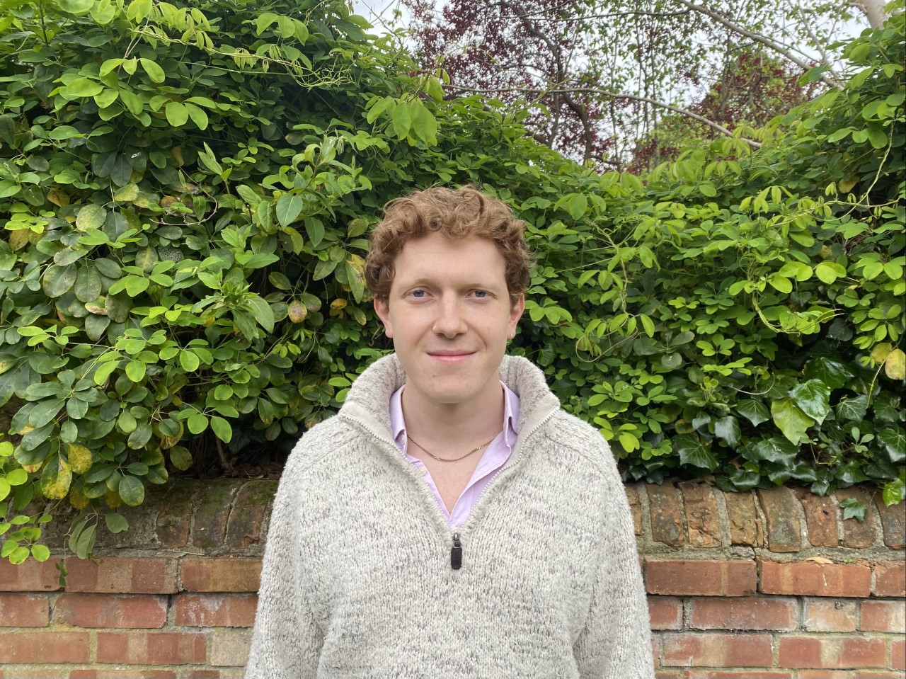 Pete Rae smiling and standing in front of bushes, wearing a cream cardigan.