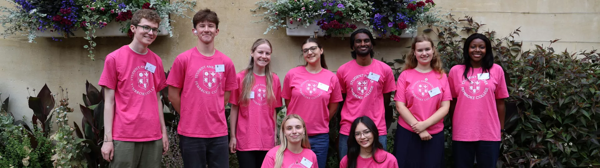 Group of sudent ambassadors in pink tshirts standing in front of wall and flowerbeds in Old Quad