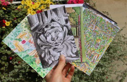 A hand holds up four colourful postcards in front of a flower box in Old Quad. The top postcard depicts a gargoyle from the tower.